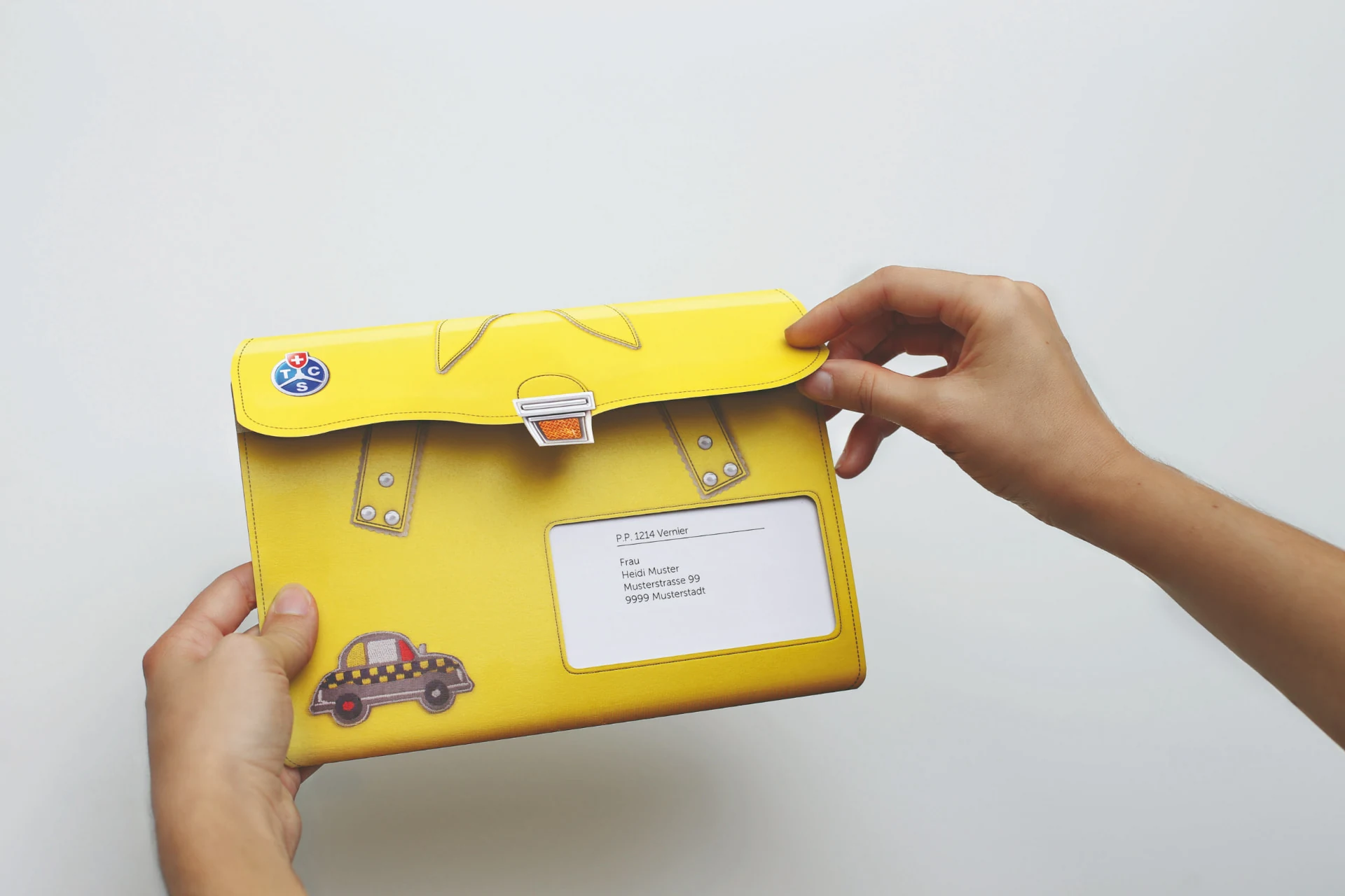 Personalisierte gelbe TCS Versandhülle im Look einer Schultasche wird von zwei Händen geöffnet.